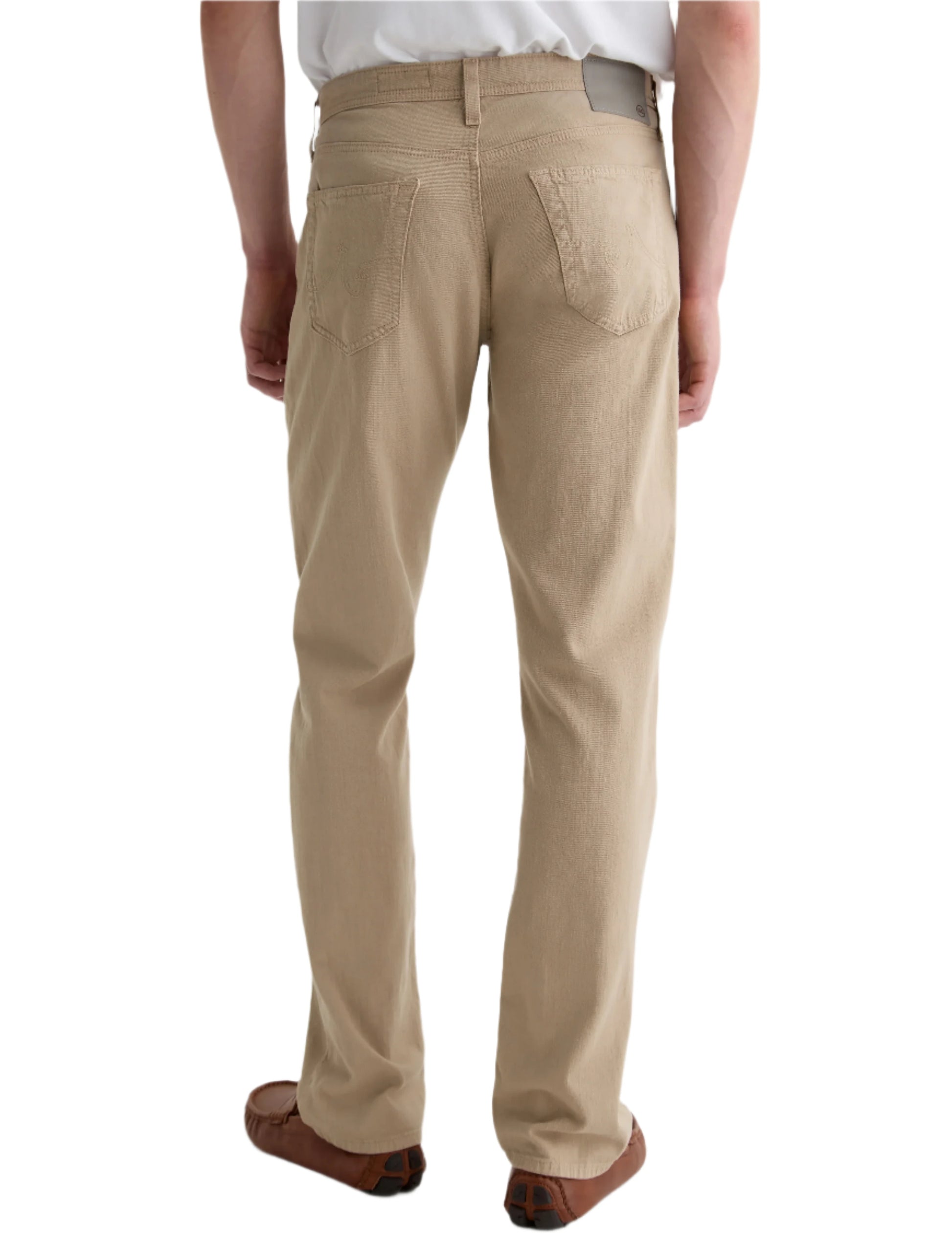 A person is shown from the back, standing against a plain white background, wearing AG Jeans' Everett Slim Straight beige jeans with a classic five-pocket design, paired with a white shirt and brown shoes.