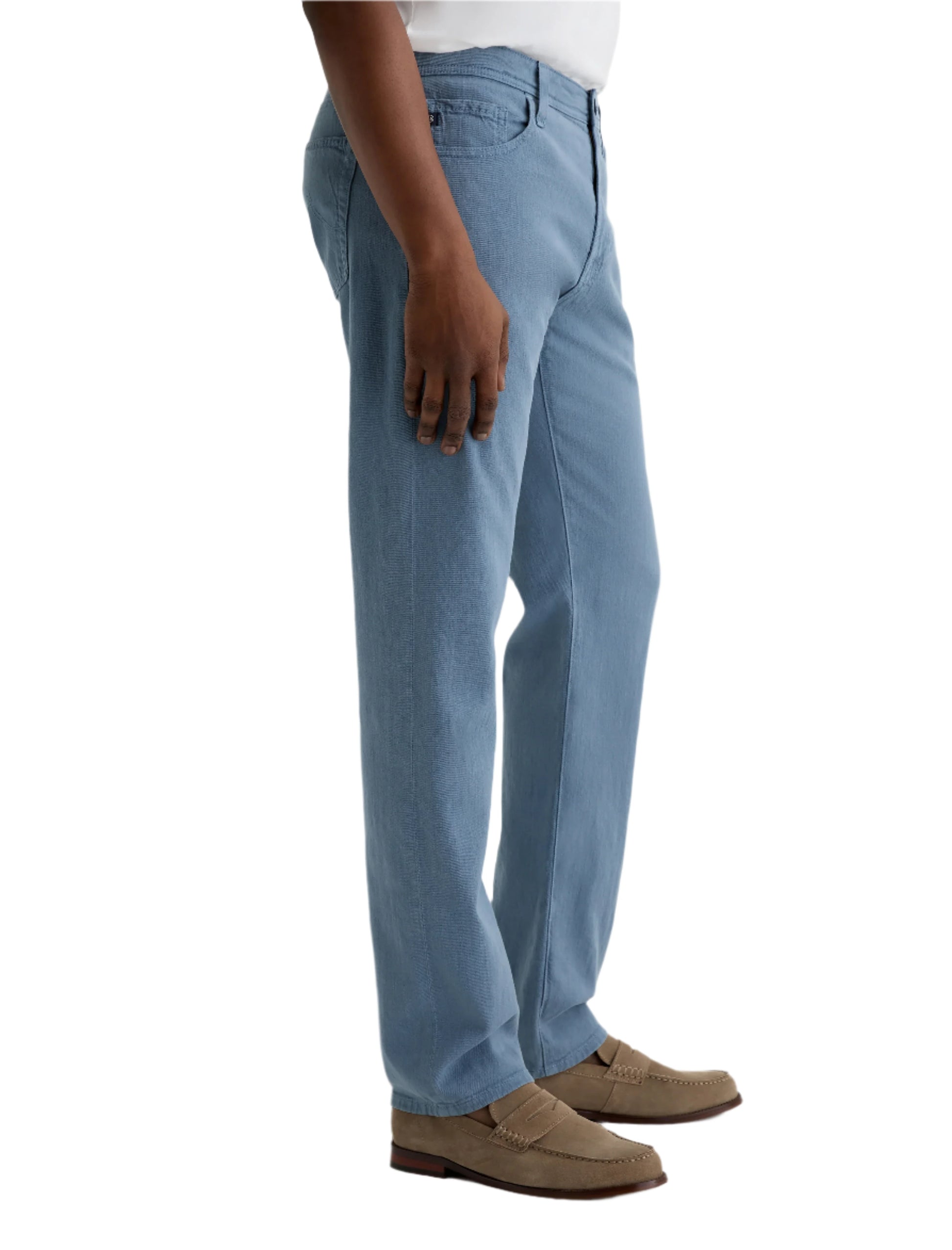 Someone wearing the Everett Slim Straight light blue jeans by AG Jeans, featuring a classic five-pocket design, paired with a white shirt and light brown loafers, stands against a plain background.