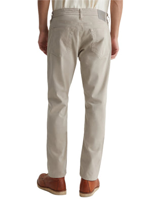 A person shown from the back is wearing AG Jeans' Everett Sueded Slim Straight pants in light beige, paired with tan leather shoes and a white shirt.