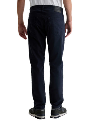 A person in a white shirt and black sneakers with white soles stands facing away, showcasing the back of the men’s AG Jeans Everett Sueded Slim Straight jeans.