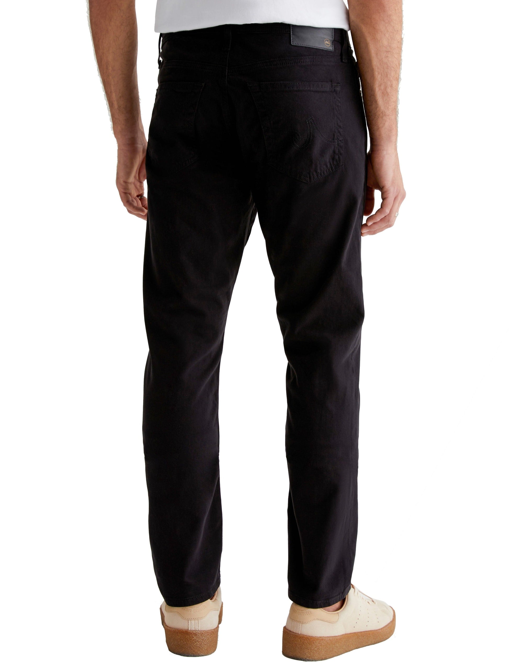 Man in black AG Jeans Everett Sueded Stretch Sateen pants and white shoes standing with a side view.