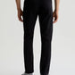 A person stands facing away from the camera, wearing AG Jeans Tellis Modern Slim Sueded Stretch Sateen men’s jeans in black, paired with grey sneakers and a dark shirt, set against a plain light background.