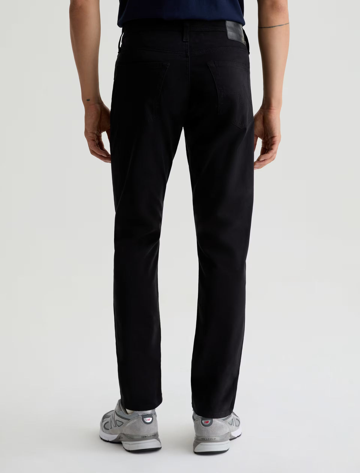 A person stands facing away from the camera, wearing AG Jeans Tellis Modern Slim Sueded Stretch Sateen men’s jeans in black, paired with grey sneakers and a dark shirt, set against a plain light background.