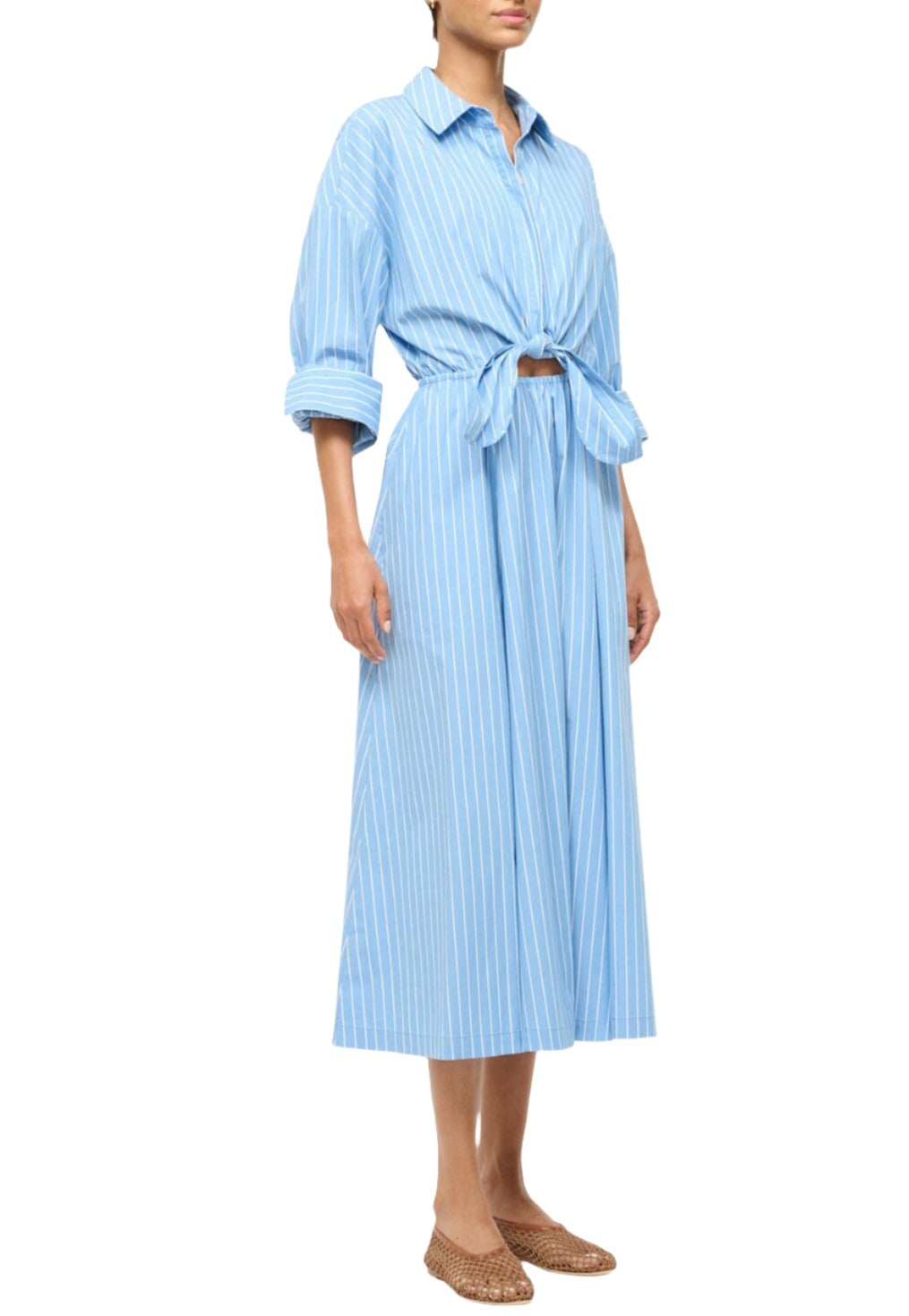 A person wearing the Staud Lisa Dress, a blue, striped cotton poplin shirtdress by Staud, featuring an elastic waist, rolled sleeves, and a tied front. The elegant midi-length dress is paired with brown woven flats.