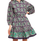 The woman is wearing a long-sleeve, knee-length Cara Cara Robin Dress in purple and green with a patterned design, featuring a subtle ruffle tier skirt for an elegant touch.