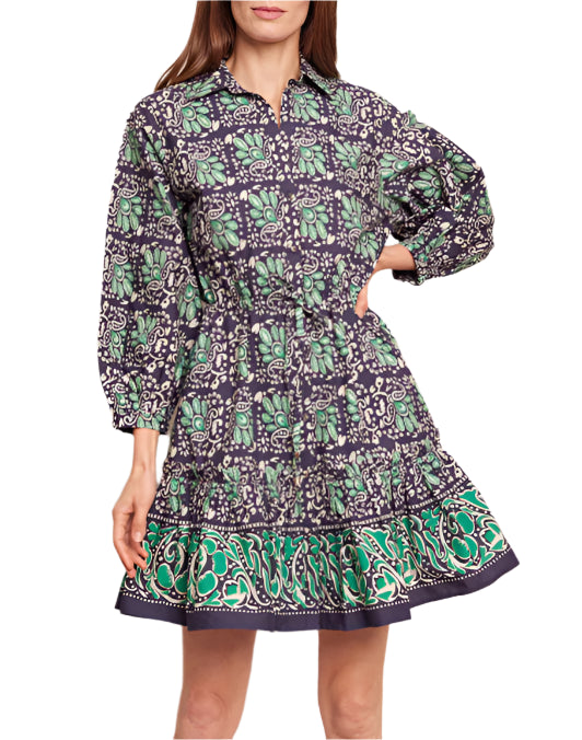 The woman is wearing a long-sleeve, knee-length Cara Cara Robin Dress in purple and green with a patterned design, featuring a subtle ruffle tier skirt for an elegant touch.