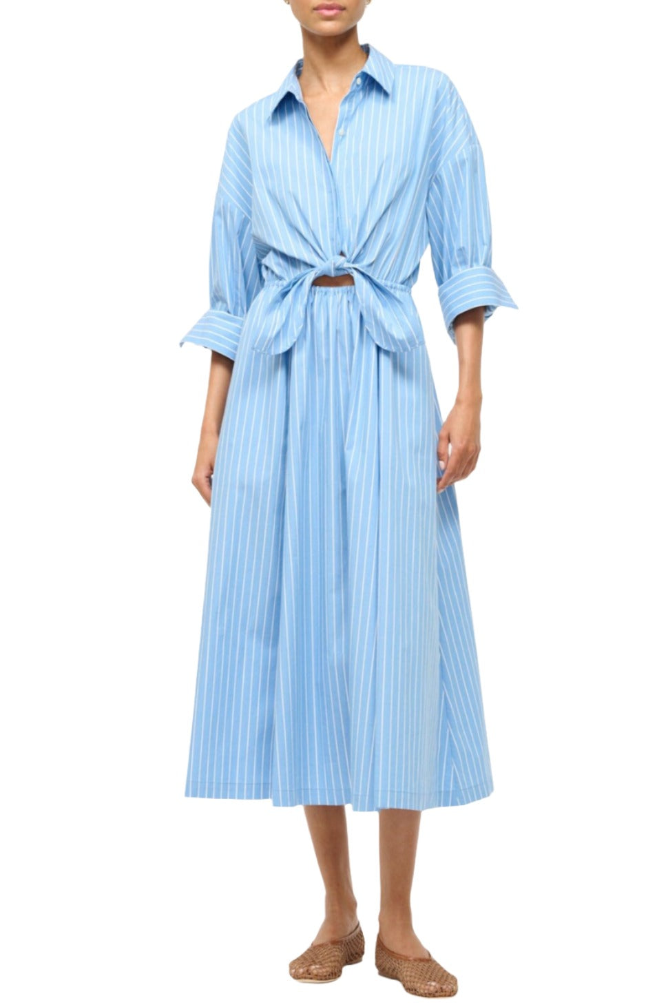 A person wearing the Staud Lisa Dress, featuring blue stripes and a midi-length design with a tie front and rolled-up sleeves, paired with woven brown slip-on shoes.