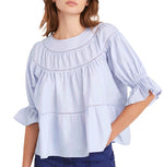 A person wearing the Merlette Sol Top, a light blue, loose-fitting Pima cotton blouse with crochet lace trim and ruffled sleeves from Merlette New York, paired with dark blue pants, stands against a plain background.