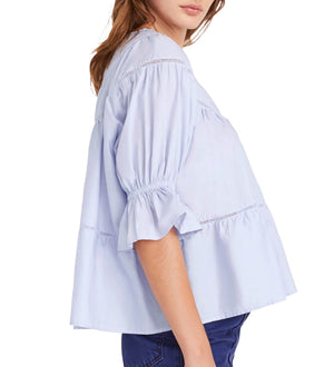 A person is wearing the Merlette Sol Top by Merlette New York, a light blue, loose-fitting blouse featuring delicate crochet lace trim, paired with blue jeans, and shown from a side angle.