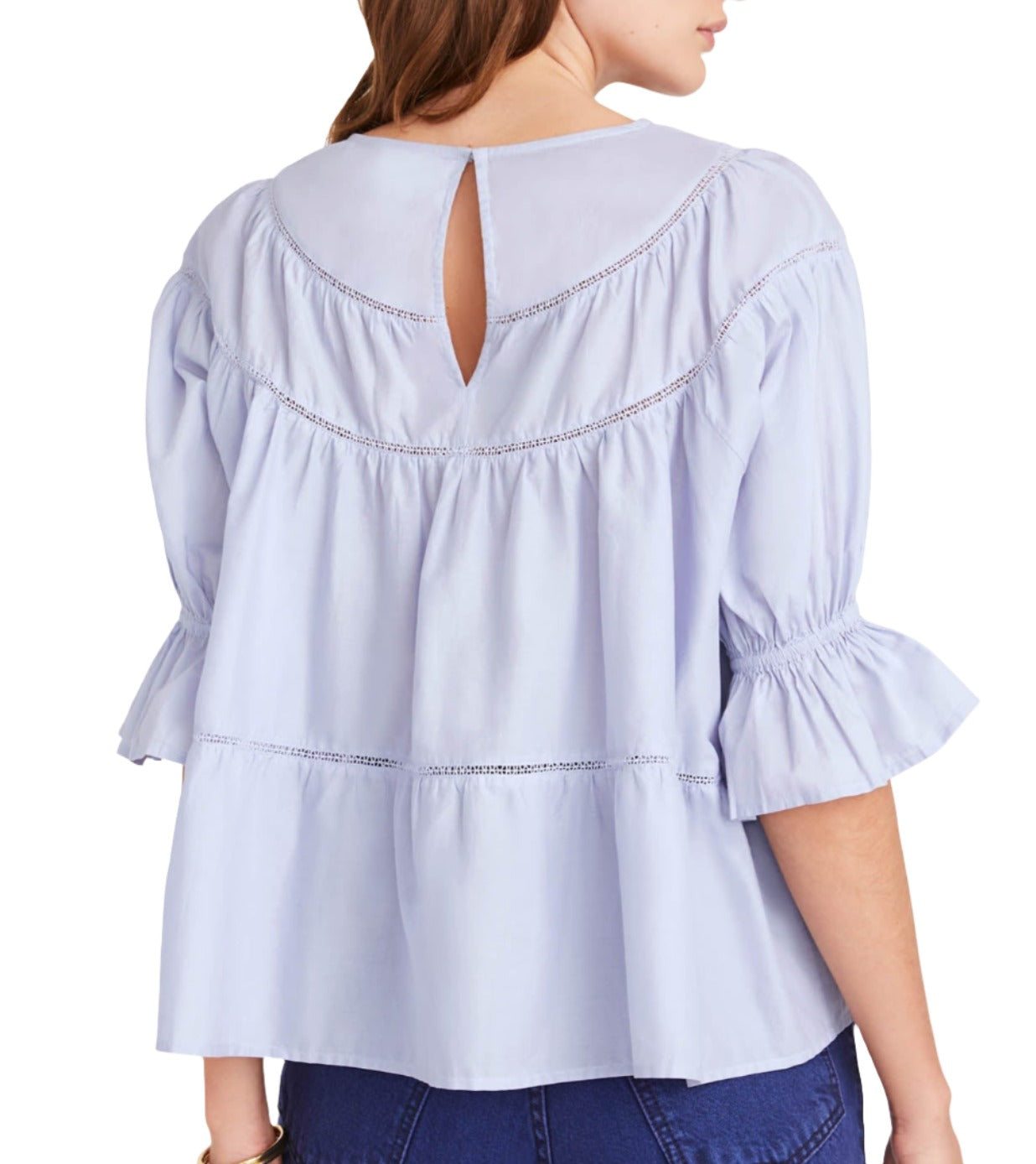 A woman is wearing the Merlette Sol Top, a light blue Pima cotton blouse by Merlette New York, featuring ruffled sleeves and a decorative back opening trimmed with crochet lace. She pairs it effortlessly with dark blue jeans.