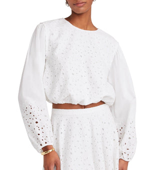 A person wearing the Merlette Linden Top from Merlette New York, a white, long-sleeve cotton eyelet blouse with bell sleeves, paired with a matching skirt.