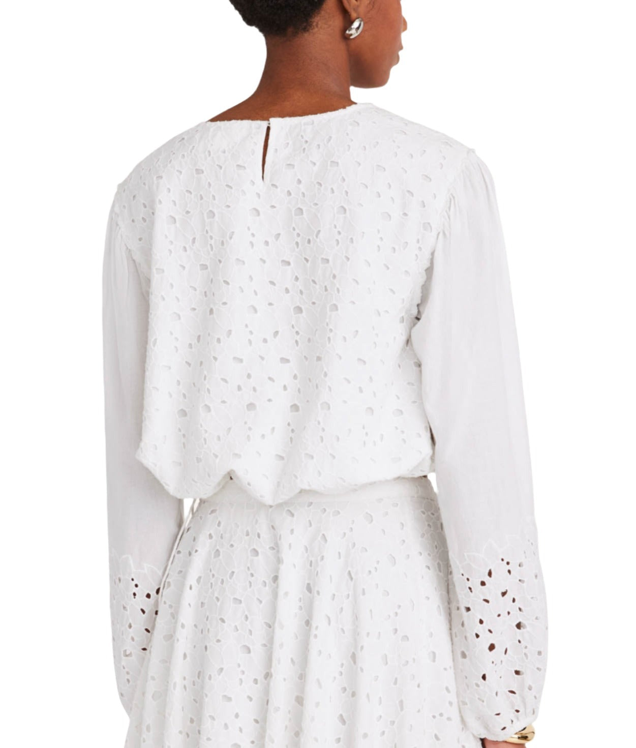 A person wearing the Merlette New York "Linden Top" in white cotton eyelet, featuring bell sleeves, paired with a matching skirt, viewed from the back.