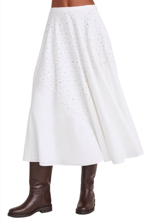 A person wearing the Merlette Merrick Skirt, a white midi skirt with cotton eyelet detailing from Merlette New York, paired with brown knee-high boots, exuding timeless elegance.