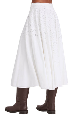 A person wearing the Merlette Merrick Skirt, a white, flowy, ankle-length skirt featuring cotton eyelet detailing from Merlette New York, paired with dark brown knee-high boots.