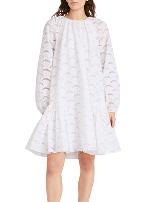 A person is wearing the Merlette Emile Dress from Merlette New York, which showcases a white, cotton silk organza design featuring a loose-fitting silhouette with a scalloped pattern and ruffled hem, highlighted by delicate crescent moon embroidery.