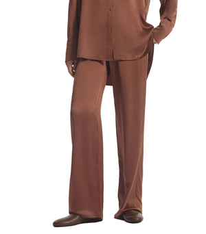 A person wearing CO’s Elastic Silk Pant—featuring a loose-fitting high-rise silhouette with an elastic waistband—paired with a matching long-sleeved top and brown shoes, standing against a plain white background.
