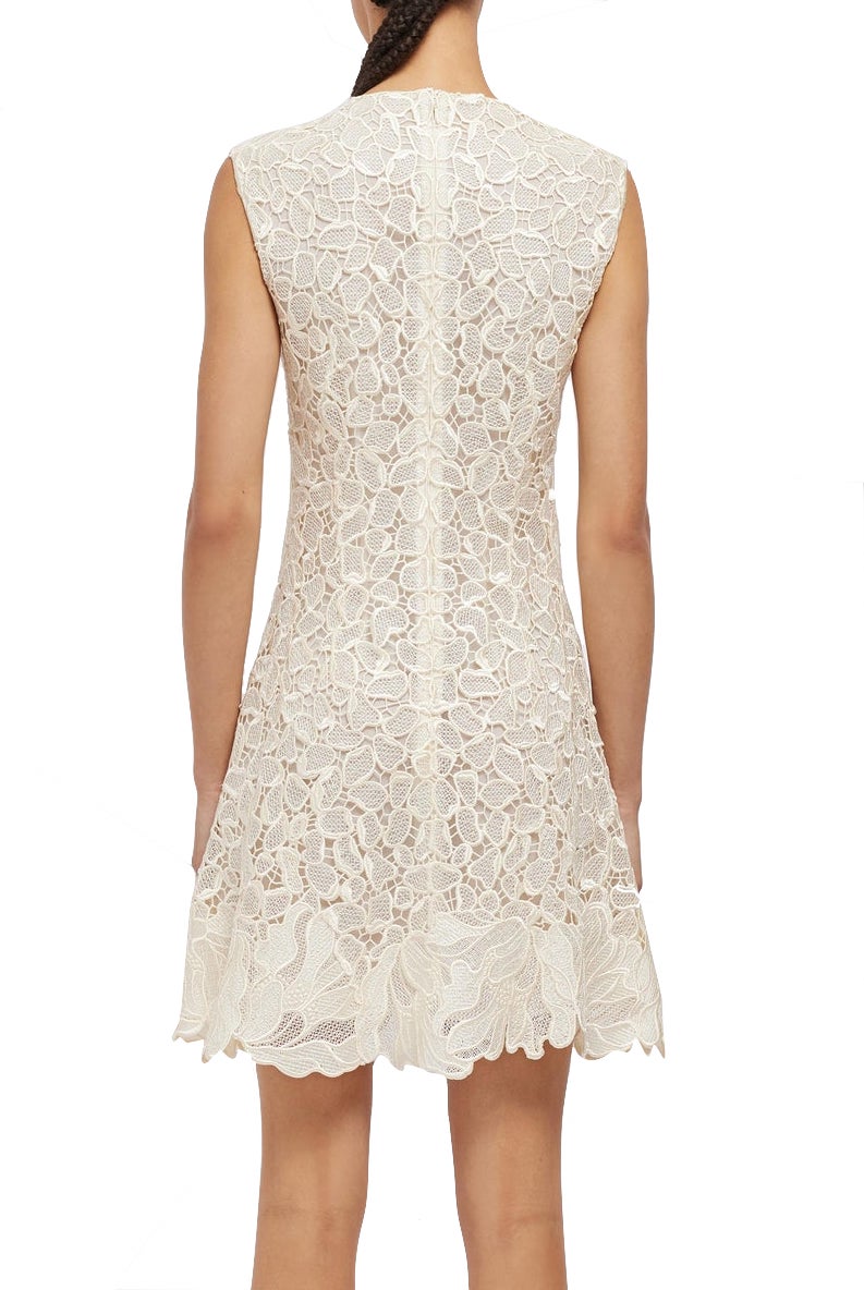 Back view of a person wearing a sleeveless, knee-length, white lace Simkhai Drake Mini Dress with floral patterns, exemplifying timeless craftsmanship by Simkhai.