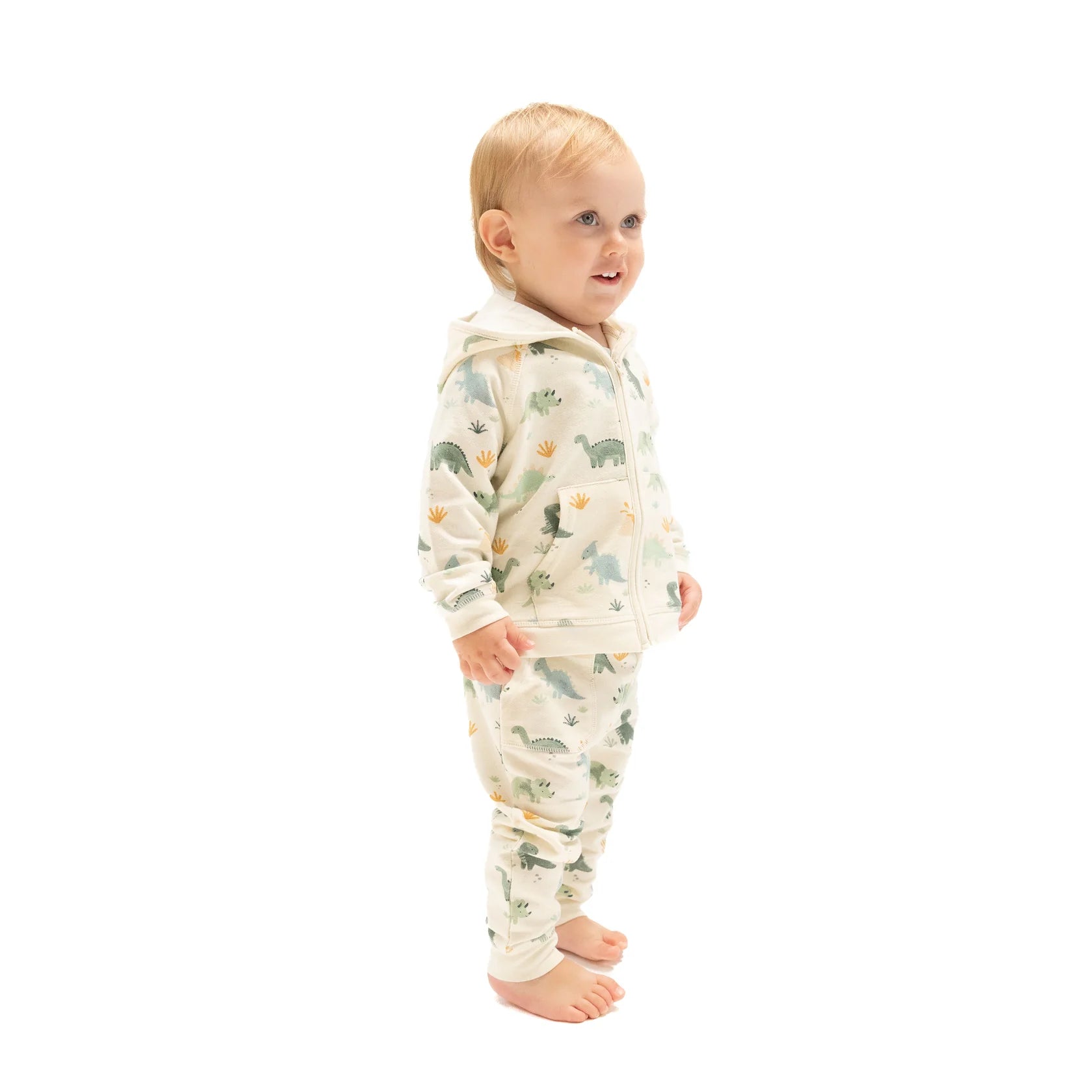 A toddler stands barefoot in the Angel Dear French Terry Zip Hoodie and Jogger Set, featuring a cream tone and playful dinosaur print, looking off to the side against a plain white background.