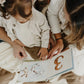 A child and two adults sit closely together, absorbed in "Hares Play - A Counting Book" by Bunnies By the Bay, intently focusing on its colorful illustrations as they gently turn the pages.