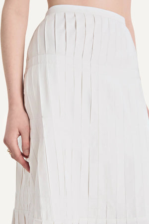 A close-up of a person wearing the Merlette New York Litus Skirt—a white, high-waisted pleated midi skirt—with their left arm resting by their side.