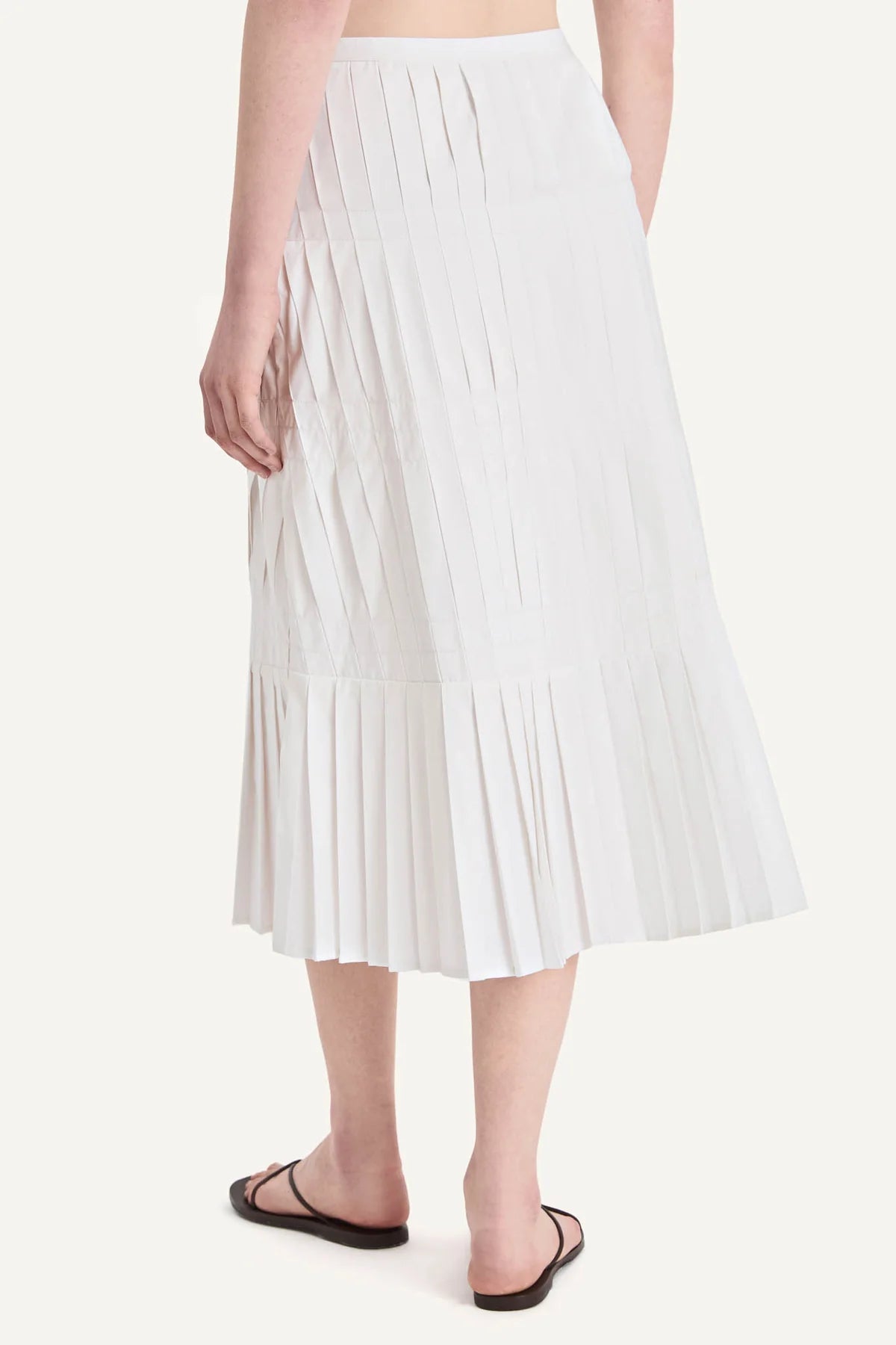 A person is seen from the back wearing the Merlette New York Litus Skirt—a white pleated midi that brings graceful movement and elegance—paired with black sandals against a plain background.
