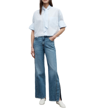 A person wearing a light blue short-sleeve shirt with a necktie detail, TWP's Tangled Up In Blue Pant—vintage washed denim wide-leg jeans with ankle slits—and white shoes, standing against a white background.