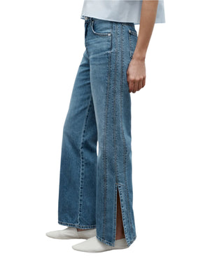 A person wearing TWP's Tangled Up In Blue Pant, vintage washed denim jeans with front slits at the hem, and white shoes is shown from the waist down in a side view.