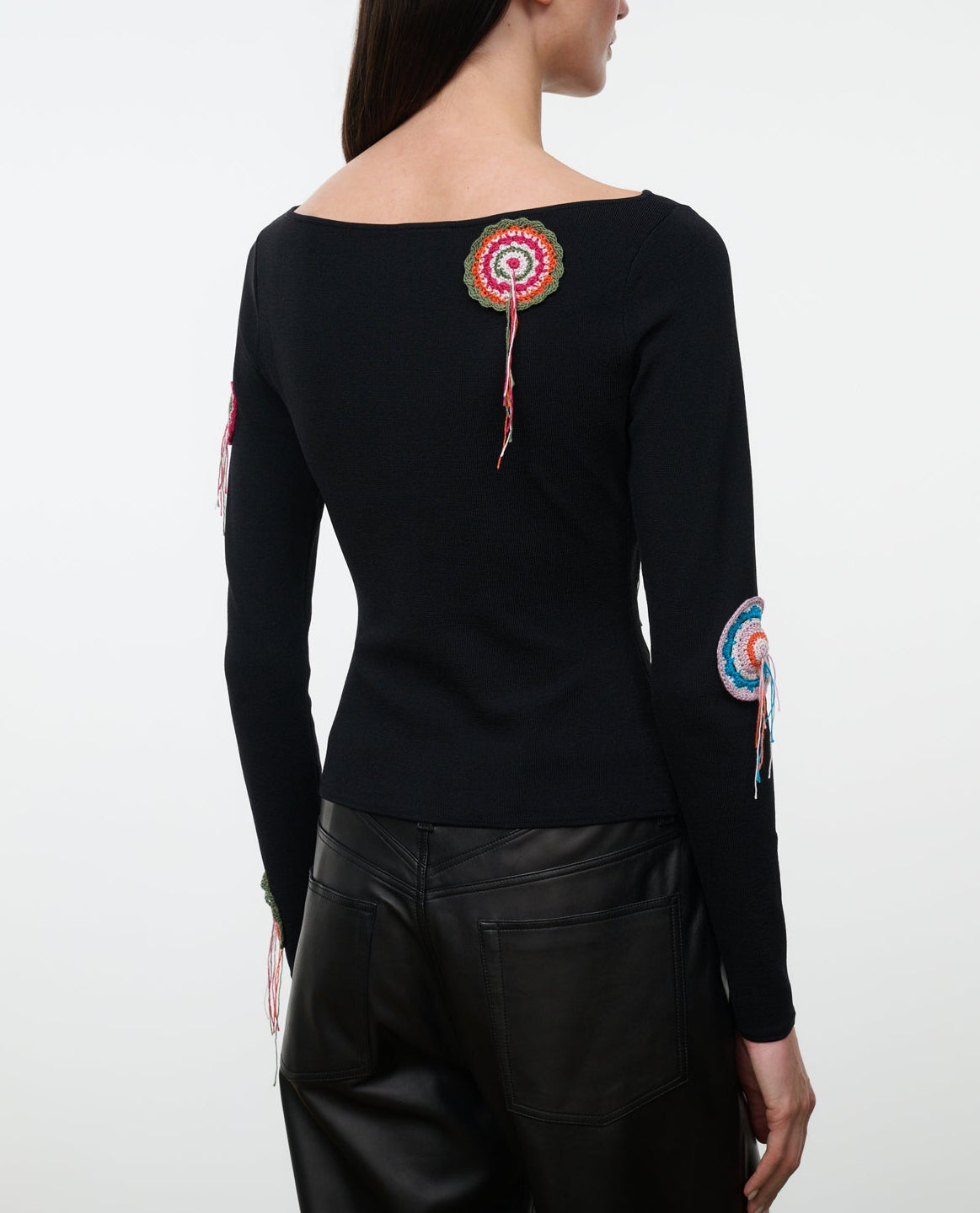 A woman wearing the Staud Rimini Top—a slim, long-sleeve style featuring a boat neckline and vibrant circular crochet appliqué patches on the back and sleeves—paired with black leather pants.