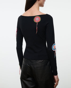 A woman wearing the Staud Rimini Top—a slim, long-sleeve style featuring a boat neckline and vibrant circular crochet appliqué patches on the back and sleeves—paired with black leather pants.
