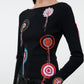 A woman wearing the Staud Rimini Top—a black slim long-sleeve shirt featuring colorful circular crochet doily appliqué—paired with black pants, shown from the chest up against a plain background.