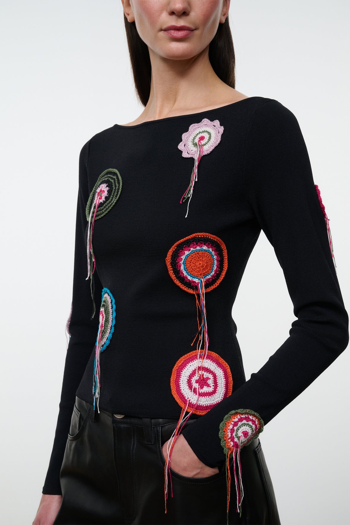 A woman wearing the Staud Rimini Top—a black slim long-sleeve shirt featuring colorful circular crochet doily appliqué—paired with black pants, shown from the chest up against a plain background.