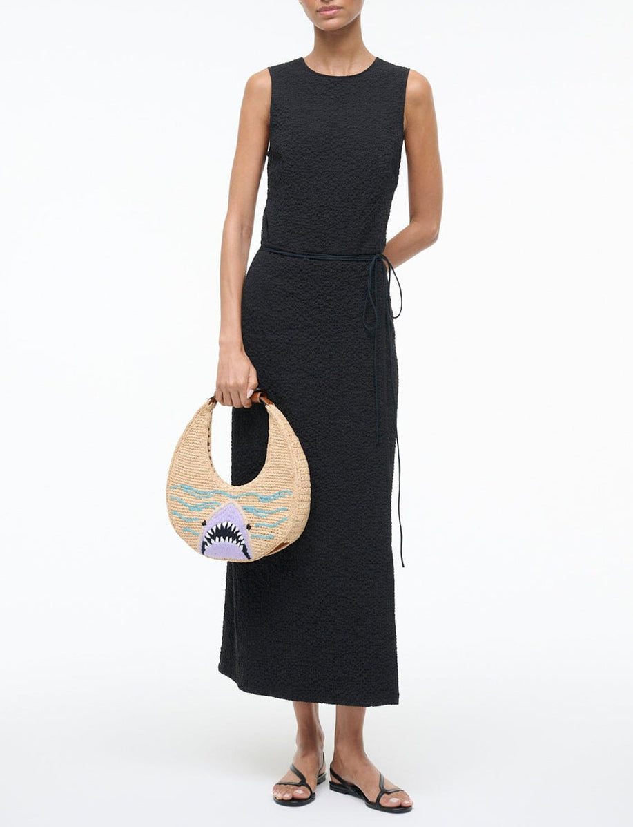 Woman wearing a sleeveless black textured maxi dress and black sandals, holding the Staud Moon Raffia Tote Bag; posed against a plain white background.