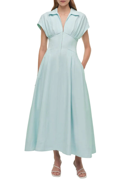 A woman wears the Simkhai Farlow Dress—a light blue cotton faille style with short sleeves, collared neckline, pleated bodice, and front seam—paired with white heeled sandals for a polished, understated look.
