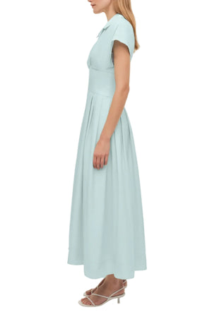 A woman stands in profile against a plain background wearing the Simkhai Farlow Dress, a light blue, collared, short-sleeve cotton faille piece with a polished, understated look, styled with white sandals.