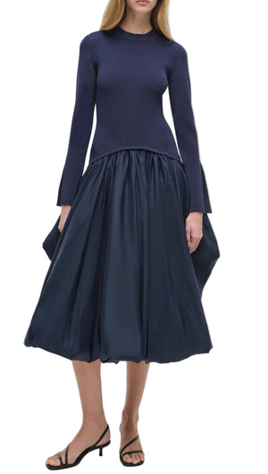 A woman in the Simkhai Kenlie Midi Dress, a navy blue mock neck long-sleeve design, styled with black open-toe sandals, exudes an elegant silhouette.
