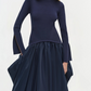 A woman stands against a plain light background wearing the Simkhai Kenlie Midi Dress, a fitted navy blue mock neck style with slit cuffs and a voluminous mid-length skirt.