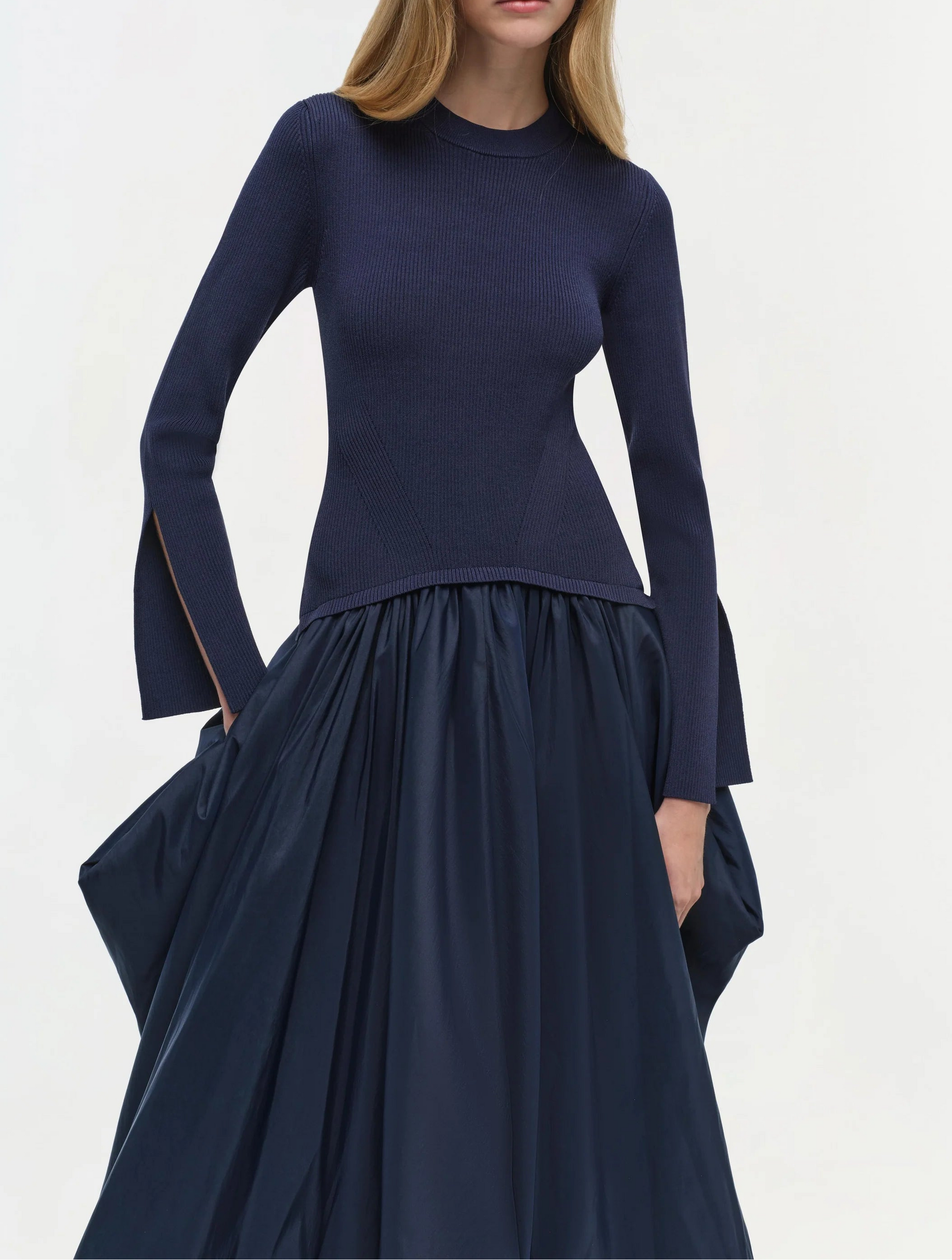 A woman stands against a plain light background wearing the Simkhai Kenlie Midi Dress, a fitted navy blue mock neck style with slit cuffs and a voluminous mid-length skirt.