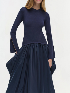 A woman stands against a plain light background wearing the Simkhai Kenlie Midi Dress, a fitted navy blue mock neck style with slit cuffs and a voluminous mid-length skirt.
