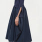 A woman stands in profile wearing the Simkhai Kenlie Midi Dress in navy with black sandals against a plain background, creating an elegant silhouette.