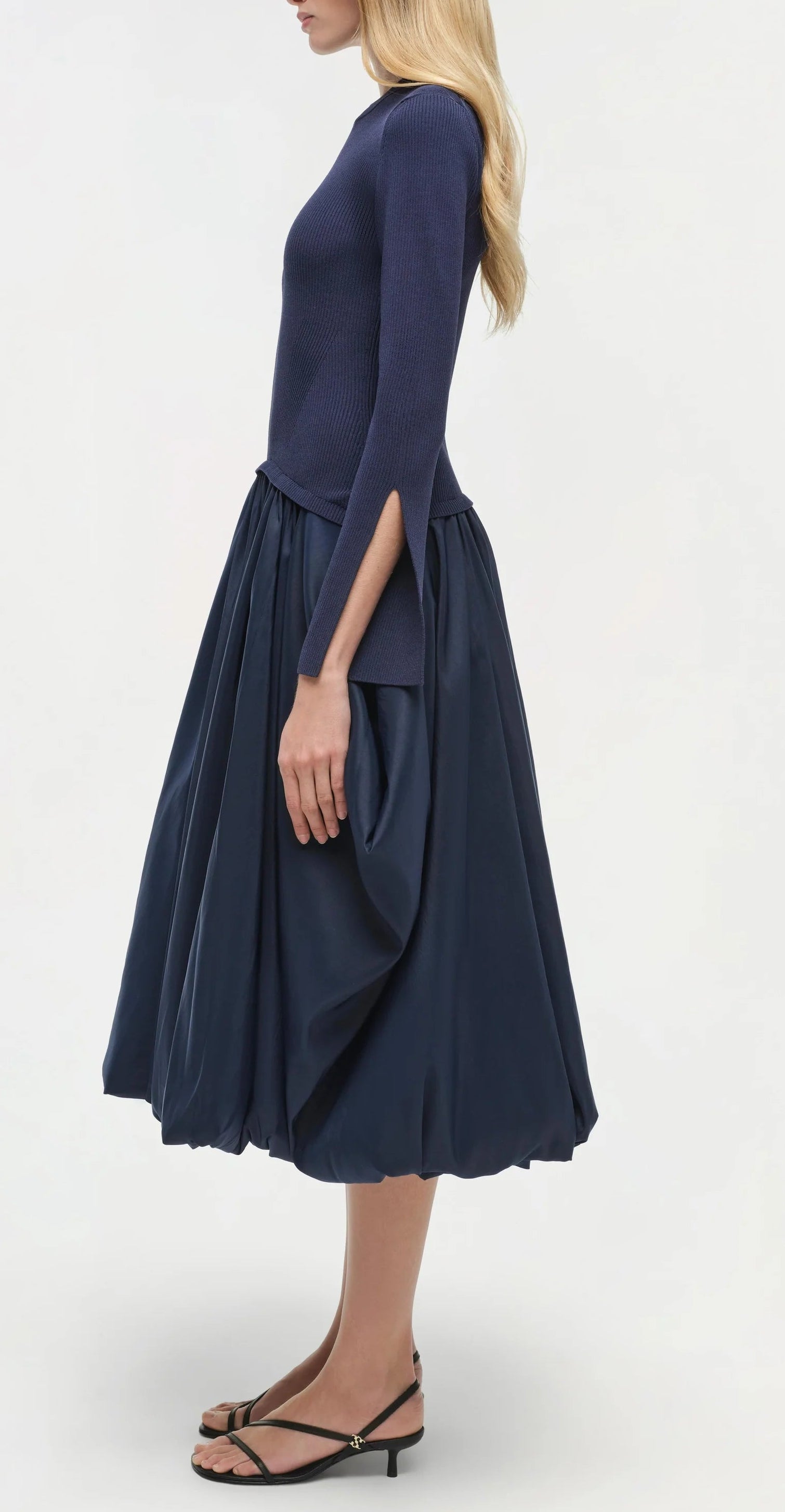 A woman stands in profile wearing the Simkhai Kenlie Midi Dress in navy with black sandals against a plain background, creating an elegant silhouette.
