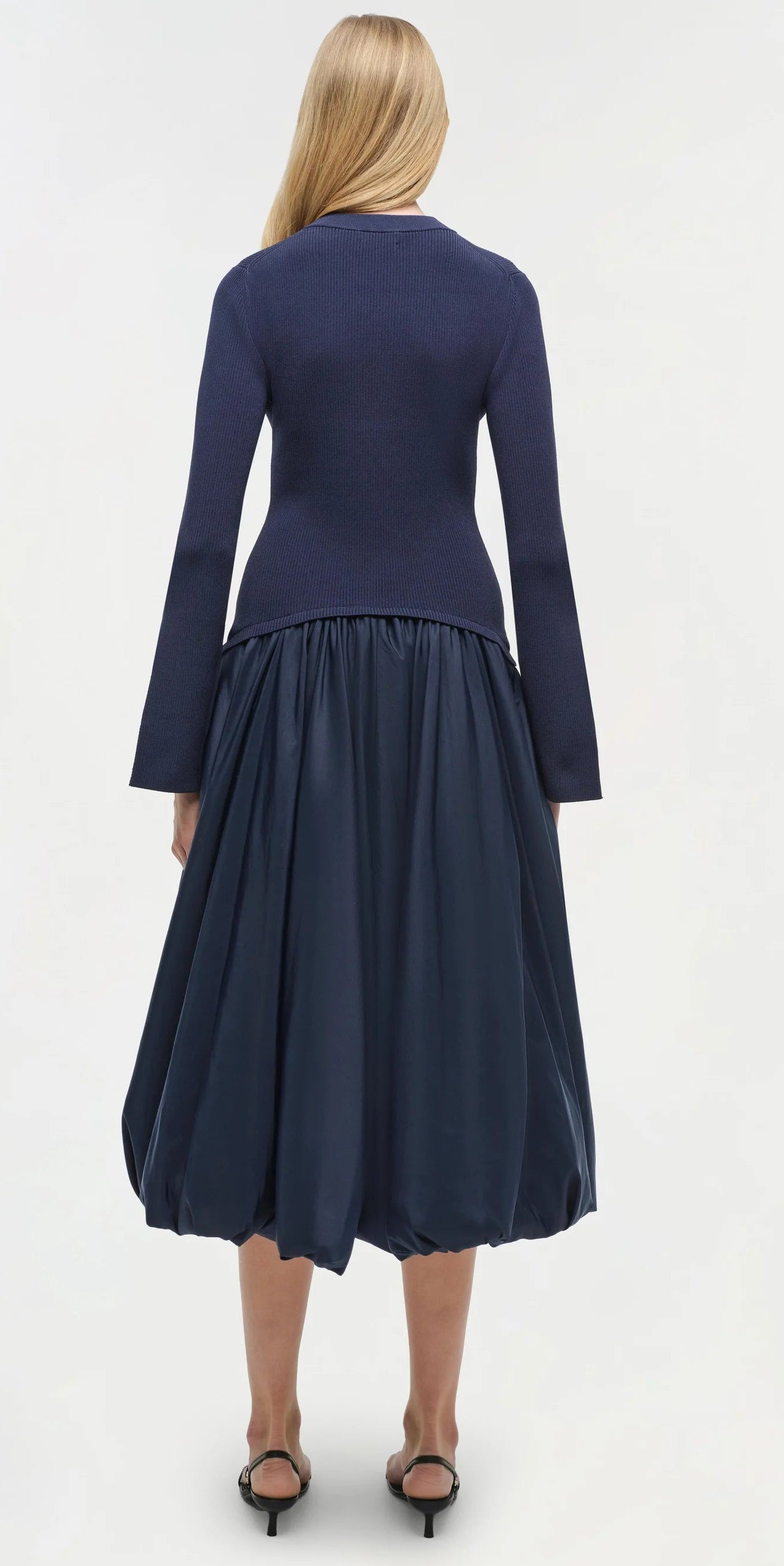 A woman stands facing away in the Simkhai Kenlie Midi Dress, styled with black sandals, against a plain light background.