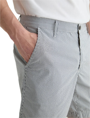 A person wearing a white shirt and AG Jeans Cipher Short blue-and-white striped pants with a mid-rise waist stands with one hand in their pocket.