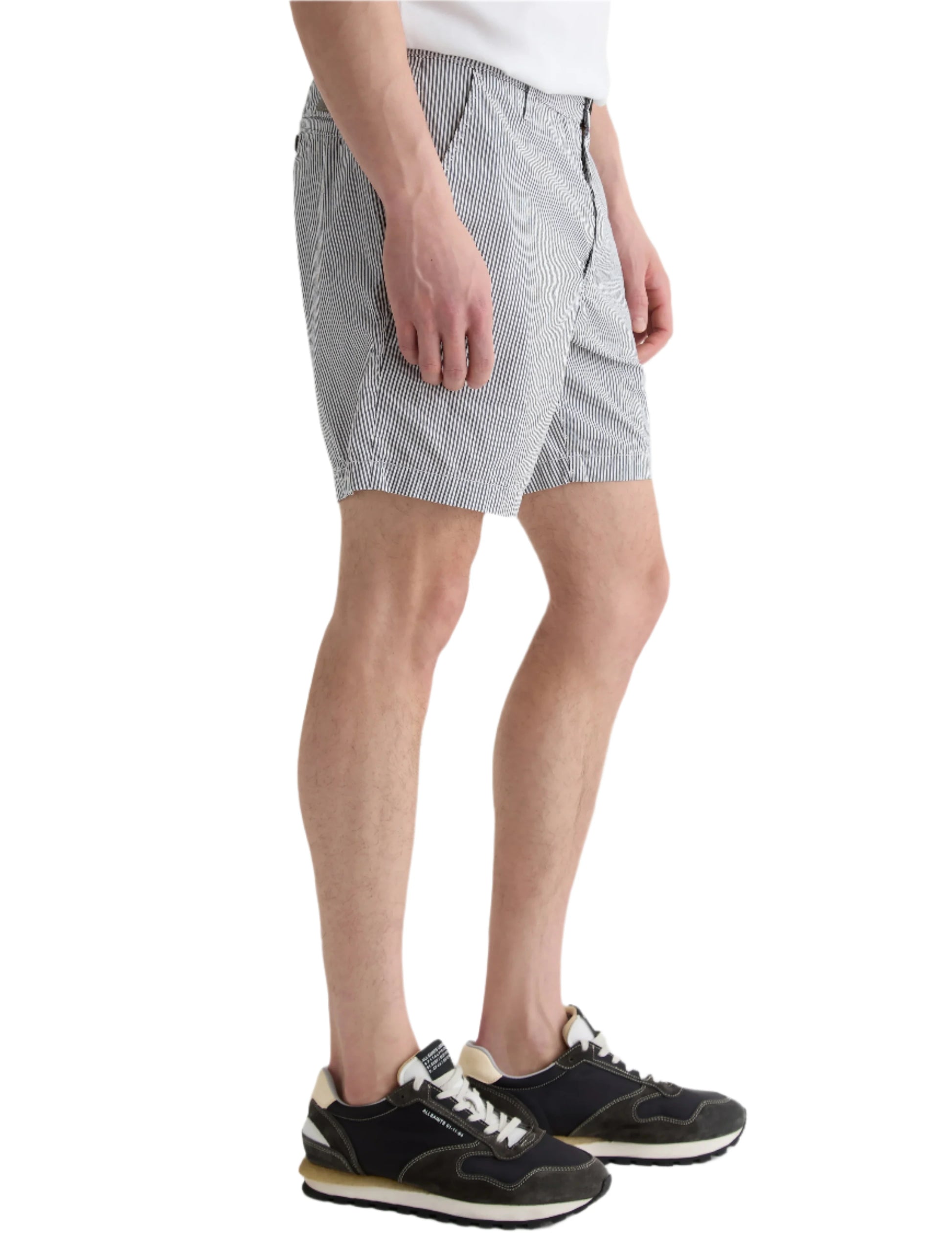 Someone wears the AG Jeans Cipher Short in white seersucker with thin vertical stripes, paired with black sneakers, standing against a white background.
