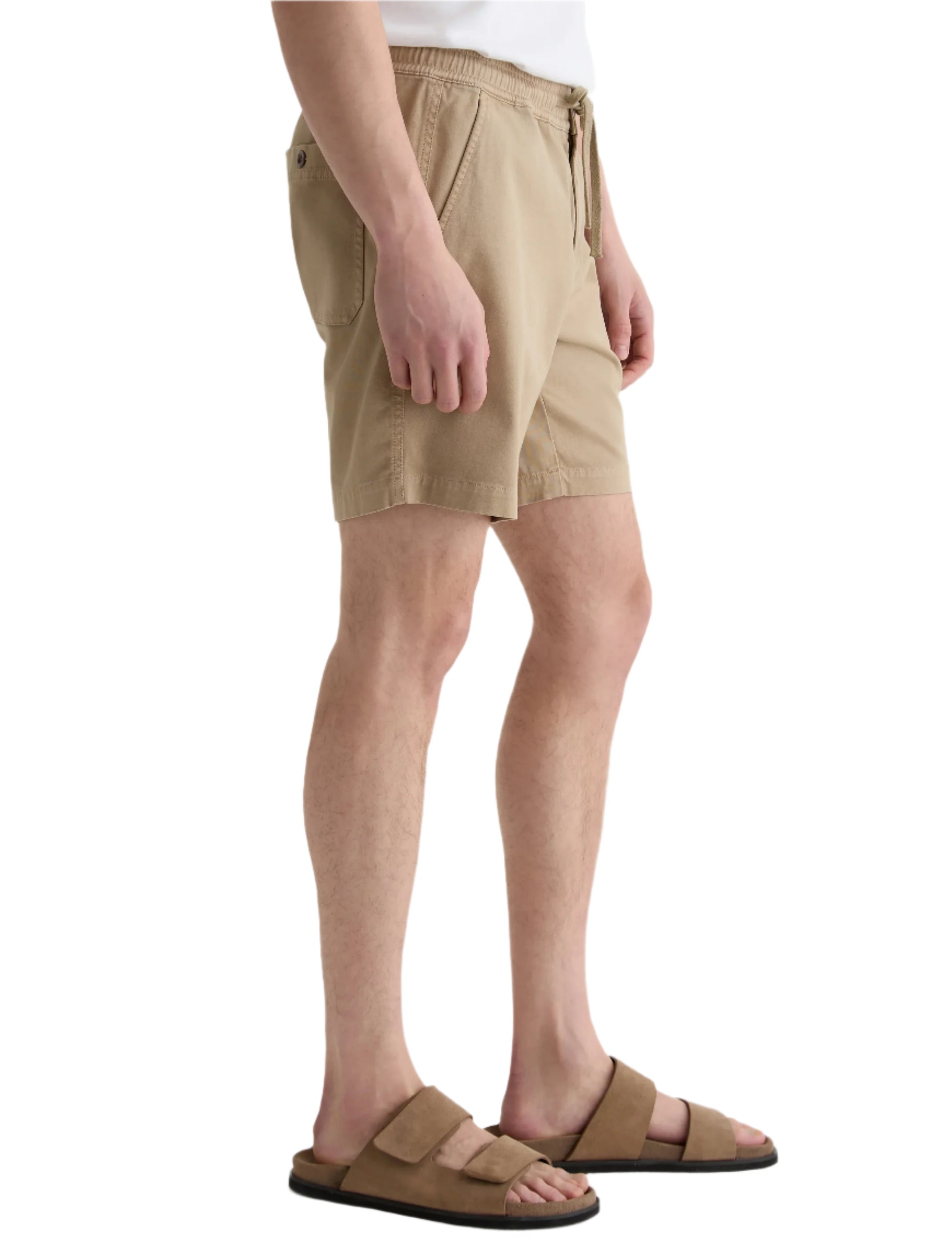 A person in AG Jeans Oliver Short beige drawstring shorts with an adjustable waist and brown sandals stands sideways against a white background.