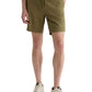 Wearing the AG Jeans Oliver Short in olive green, made from a breathable twill blend with an adjustable drawstring waist, styled with a white shirt and beige sneakers; one hand in pocket, standing against a white background.