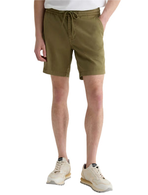 Wearing the AG Jeans Oliver Short in olive green, made from a breathable twill blend with an adjustable drawstring waist, styled with a white shirt and beige sneakers; one hand in pocket, standing against a white background.