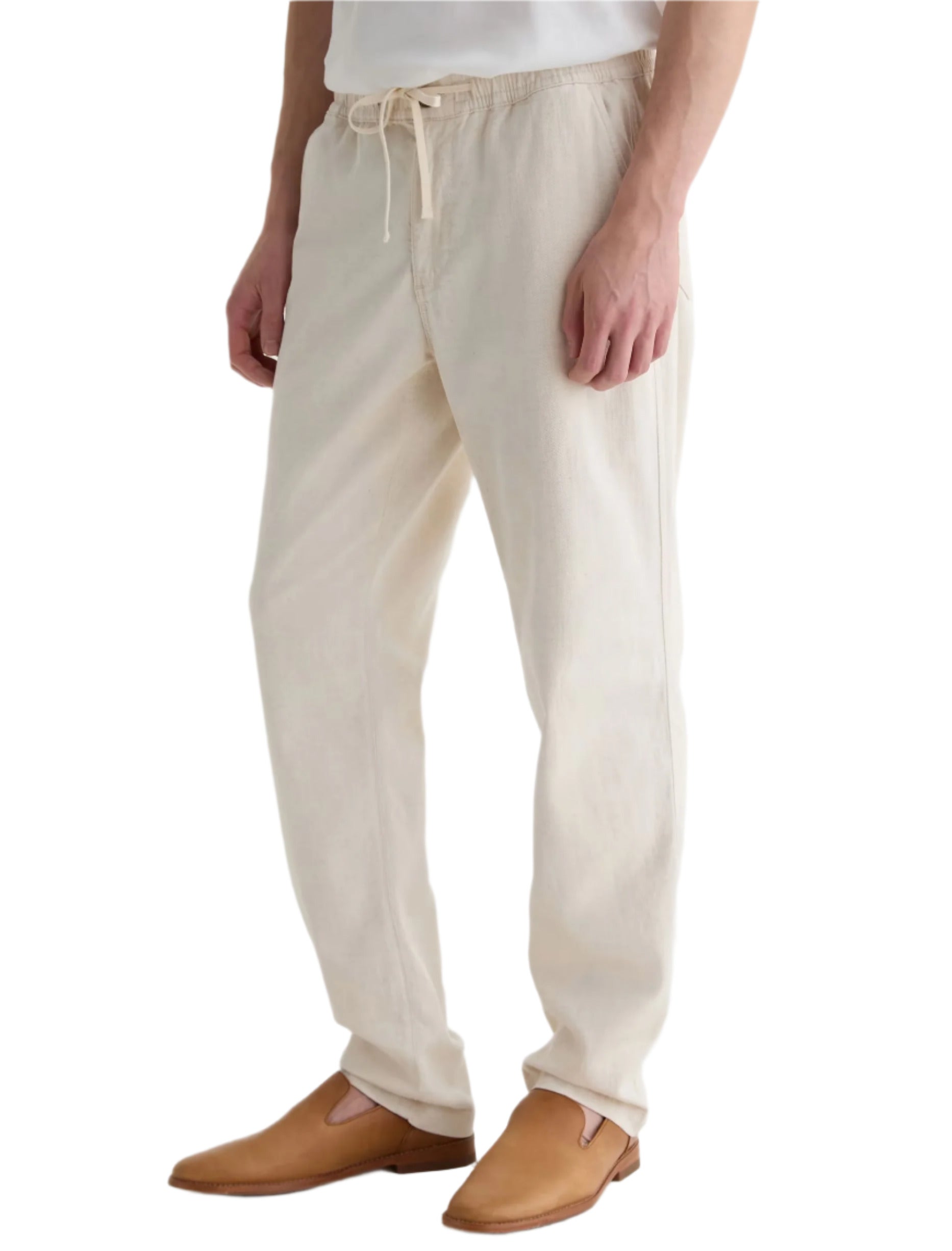 A person wears AG Jeans' Tate Drawstring Pant in light beige, paired with a white shirt and tan slip-on shoes, shown from the waist down against a white background.