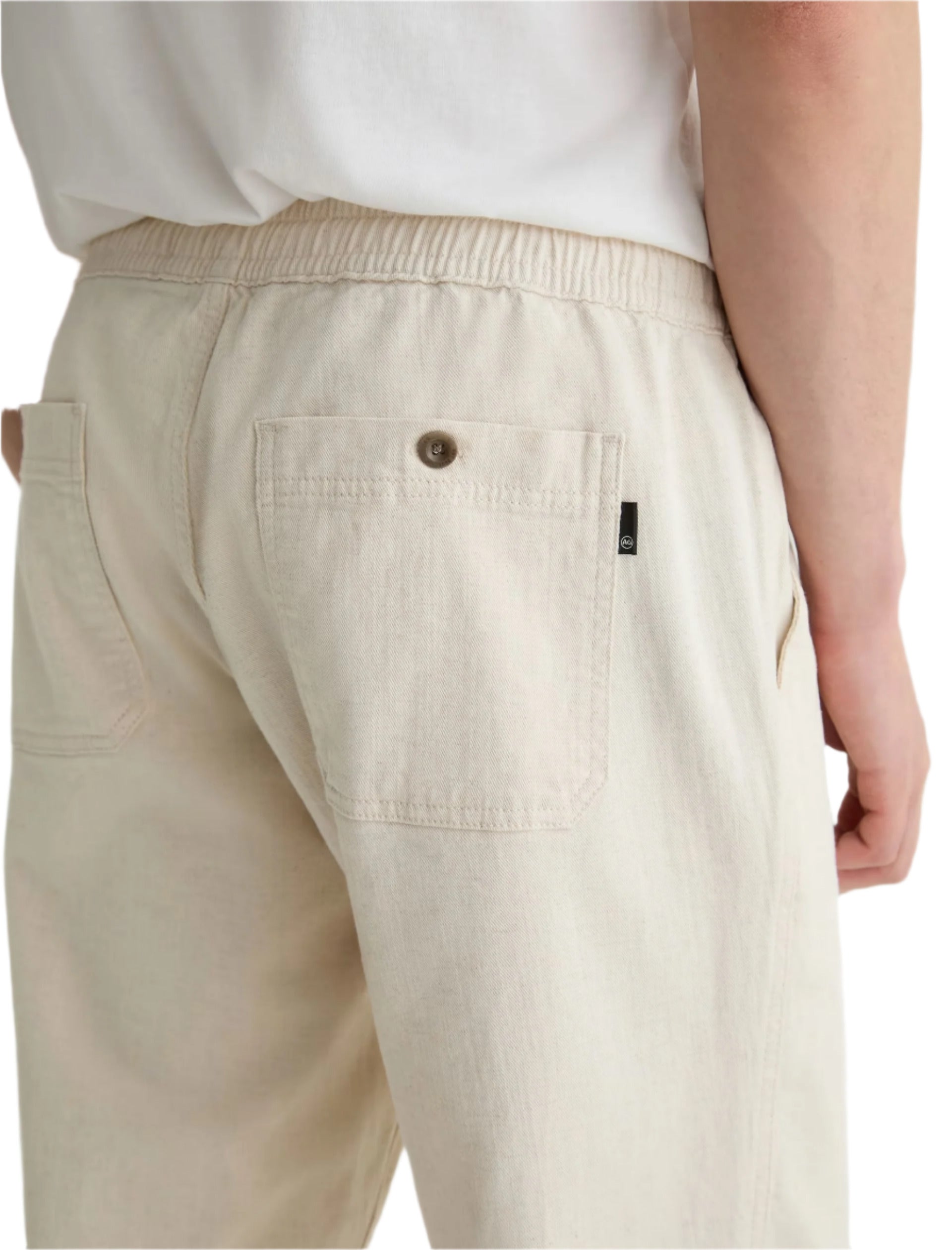 A person wearing the Tate Drawstring Pant by AG Jeans, made from light beige twill linen with patch pockets and an elastic waistband, shown from the back and cropped at the waist.