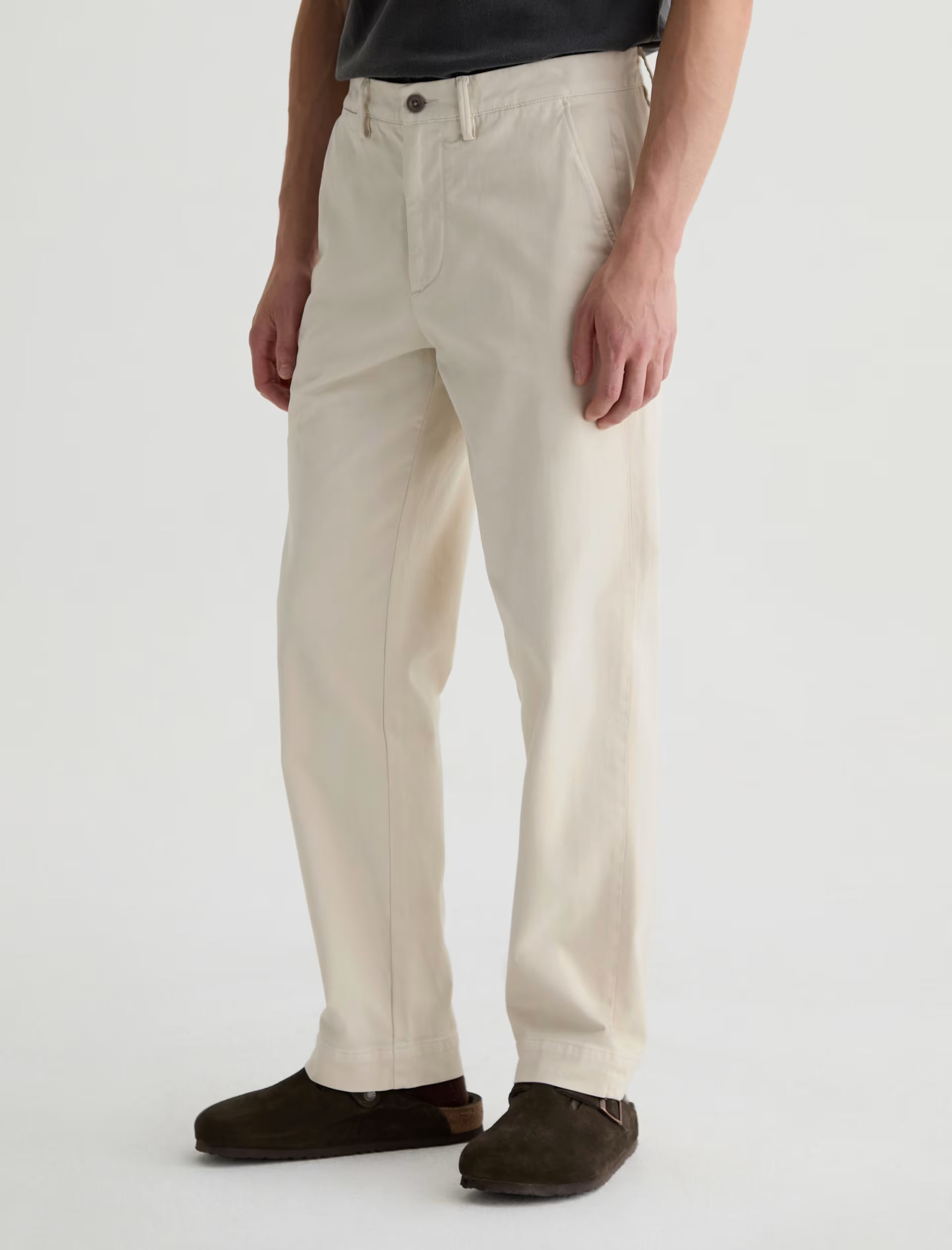 Wearing the Archie Sueded Trouser by AG Jeans in a cream shade with a relaxed straight-leg fit, paired with brown suede shoes, the individual stands against a plain light background.