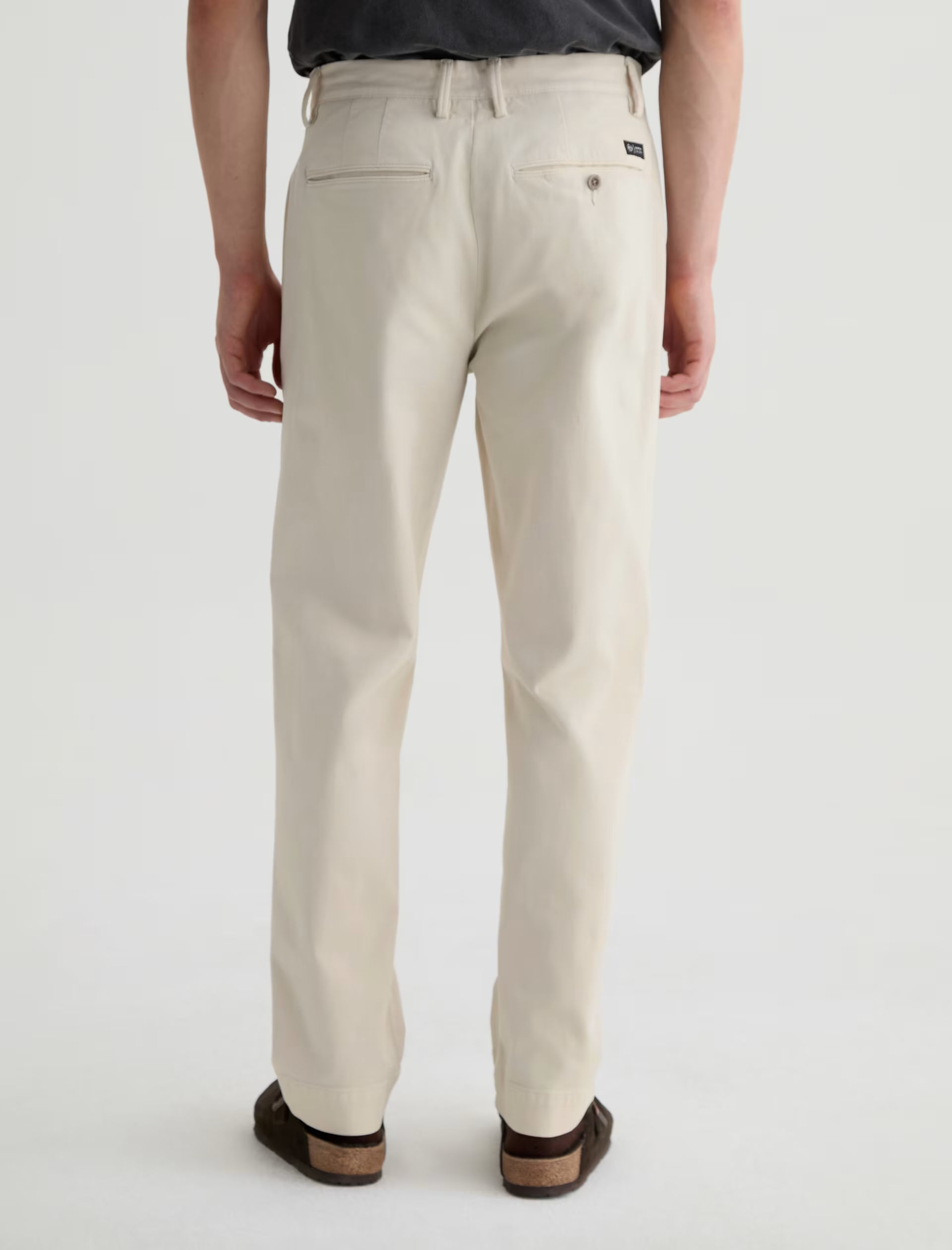 A person is shown from the back against a plain, light background wearing the Archie Sueded Trouser by AG Jeans in a light beige khaki color, paired with a dark shirt and brown shoes.